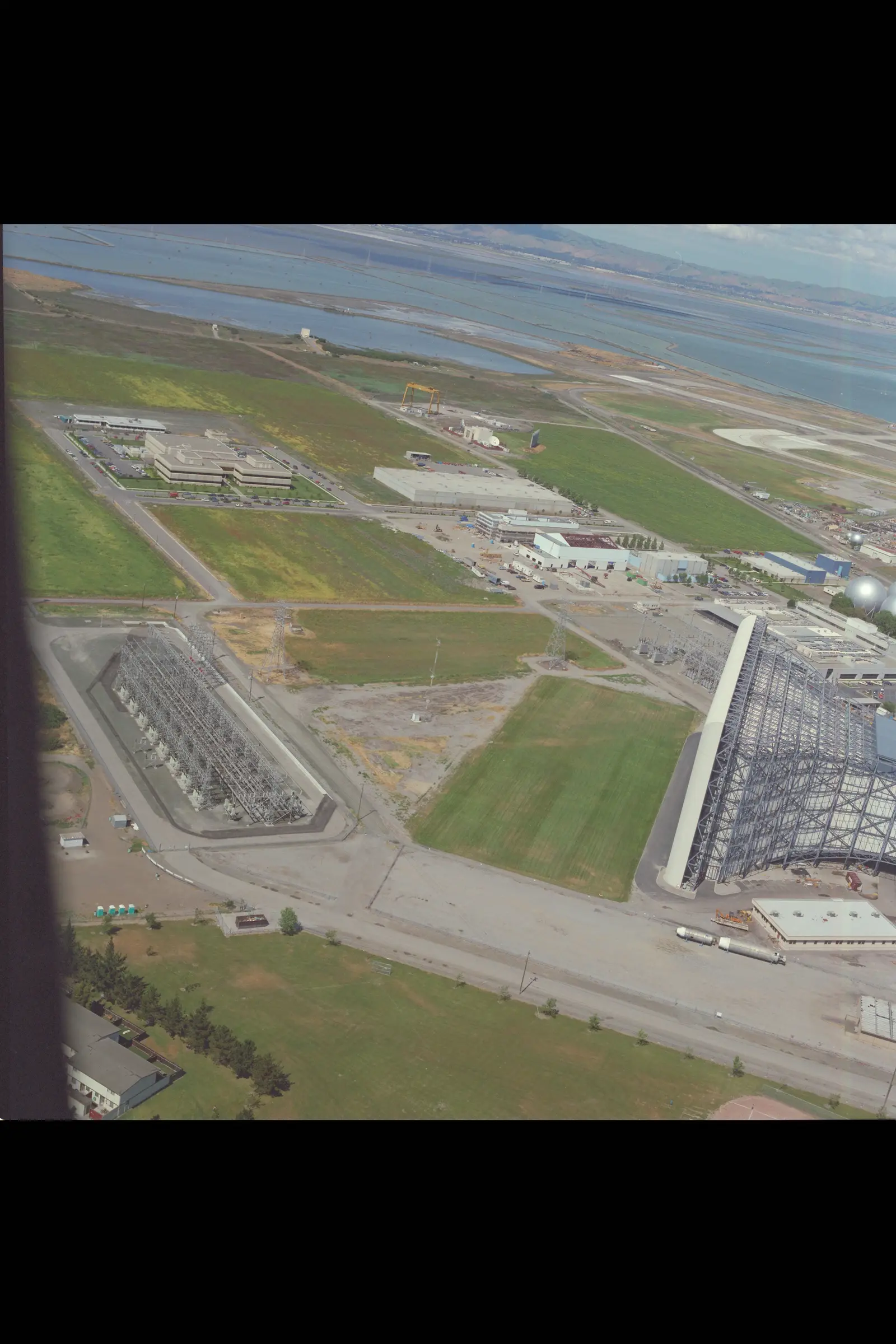 An aerial photograph shows a large industrial facility featuring a prominent electrical substation on the left and a massive, partially enclosed structure on the right, situated near a body of water.