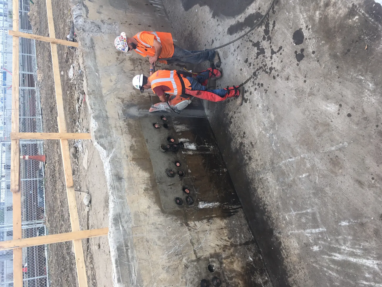 Two construction workers wearing hard hats and high-visibility vests are using equipment in a concrete substation basement lined with pipes and valves. The photographer titled it “Demolition of an old power substation basement. This substation has been replaced by a new power substation constructed as part of East Side Access. 10-07-2019 (48882438092)”.