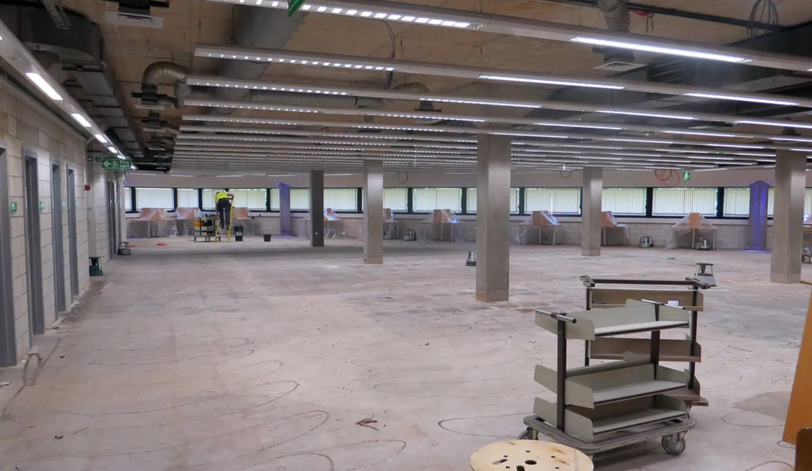 A wide, empty room under construction with exposed ceiling pipes, concrete floors, and a worker in the background. The photographer titled it “02 Inside - during - 02-08-13”.