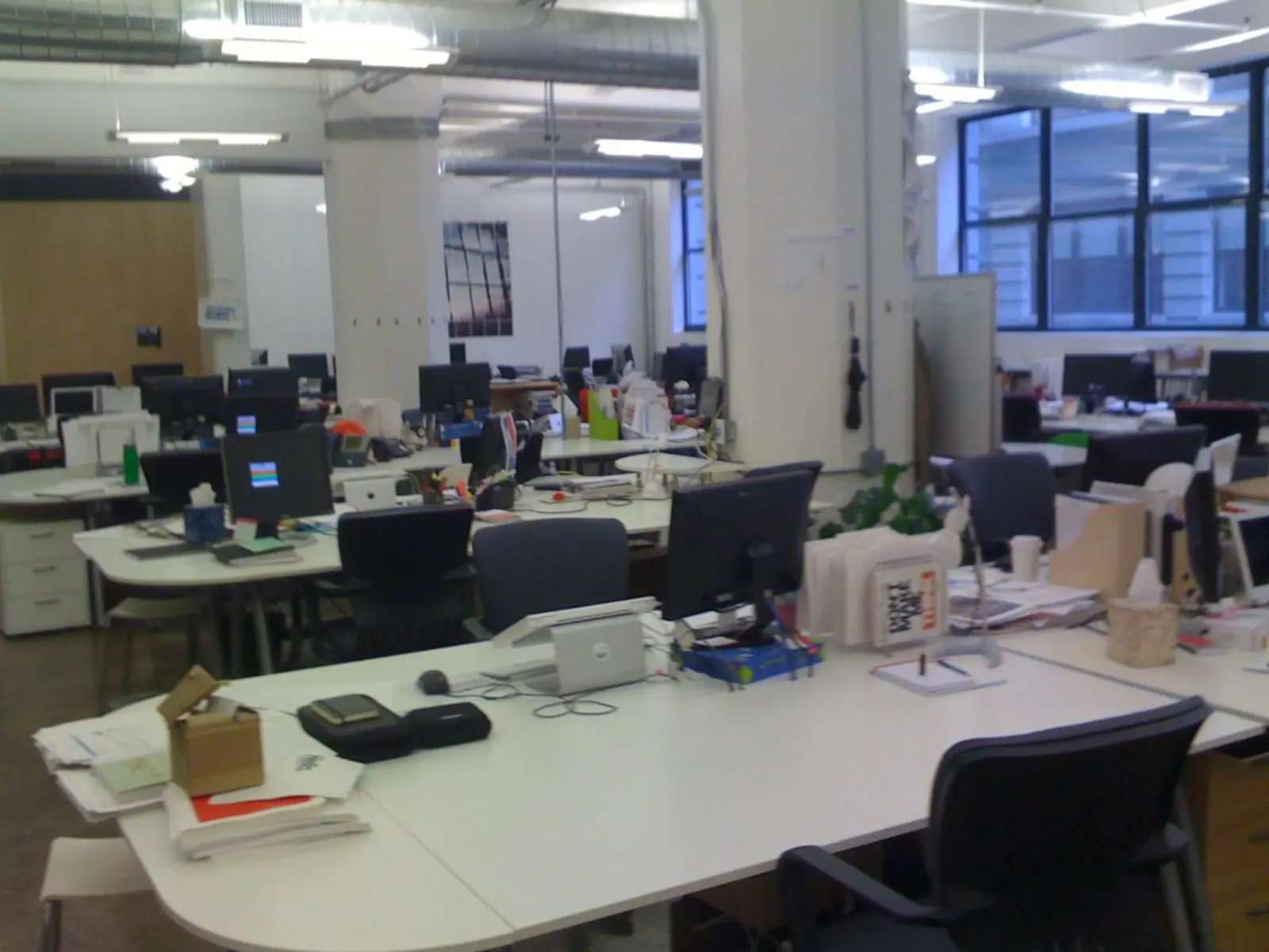 An open-plan office space features multiple white desks equipped with computer monitors, black office chairs, and scattered paperwork, with an open cardboard box resting on a desk in the foreground.